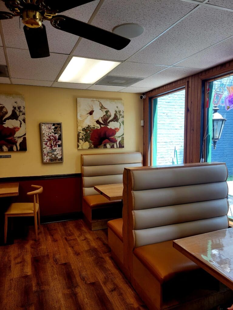 An empty, well-lit restaurant dining room with booth seating, warm yellow walls, wooden flooring, and decorative floral artwork.