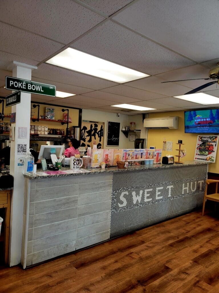An interior view of a candy or sweet shop counter with a 'Sweet Hut' sign, displaying various confections and products behind a gray counter in a warmly-lit retail space.