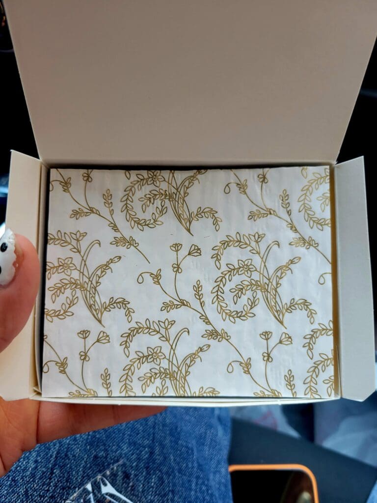 An overhead view of a person holding open a white box containing cream and gold floral patterned wallpaper or decorative paper.