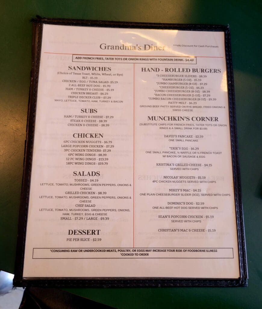 A framed restaurant menu displayed indoors listing sandwiches, hand-rolled burgers, chicken, salads, desserts, and other dining options.