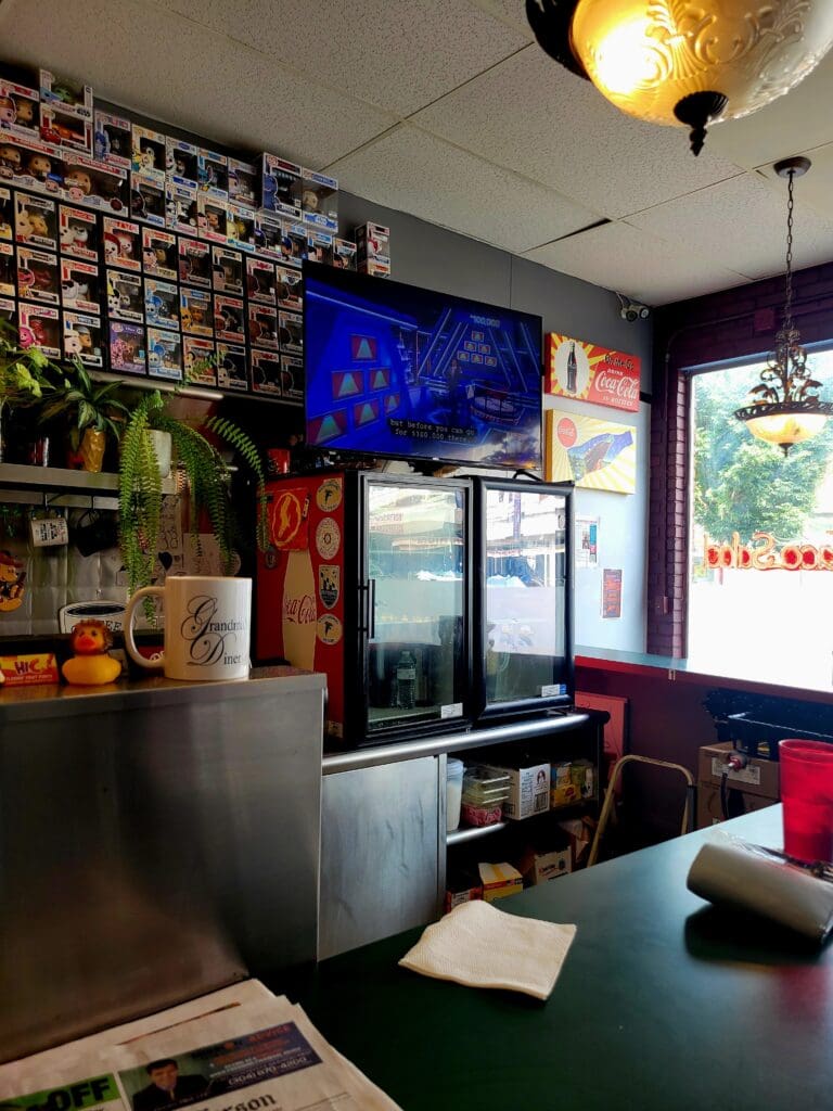 An interior view of a casual food establishment or café featuring a service counter with refrigerated displays, eclectic wall decorations, and vibrant purple and teal accents throughout the space.