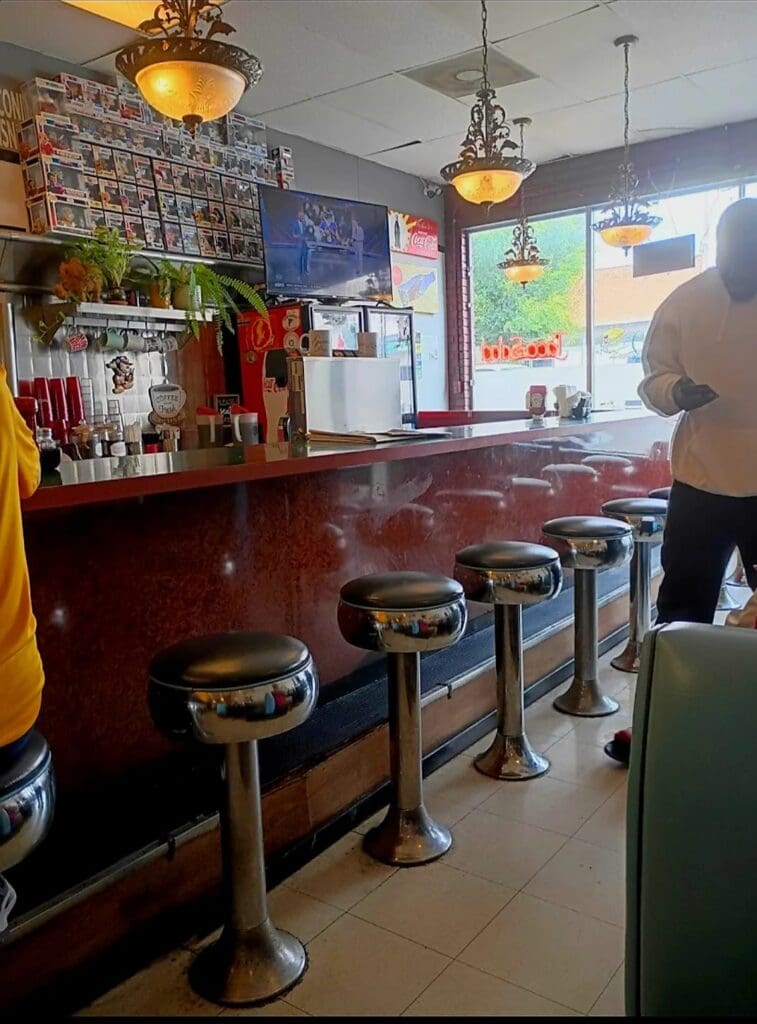 A classic retro diner interior with a red counter, chrome bar stools, and vintage decorations including neon signs on blue walls, with one person visible in the background.