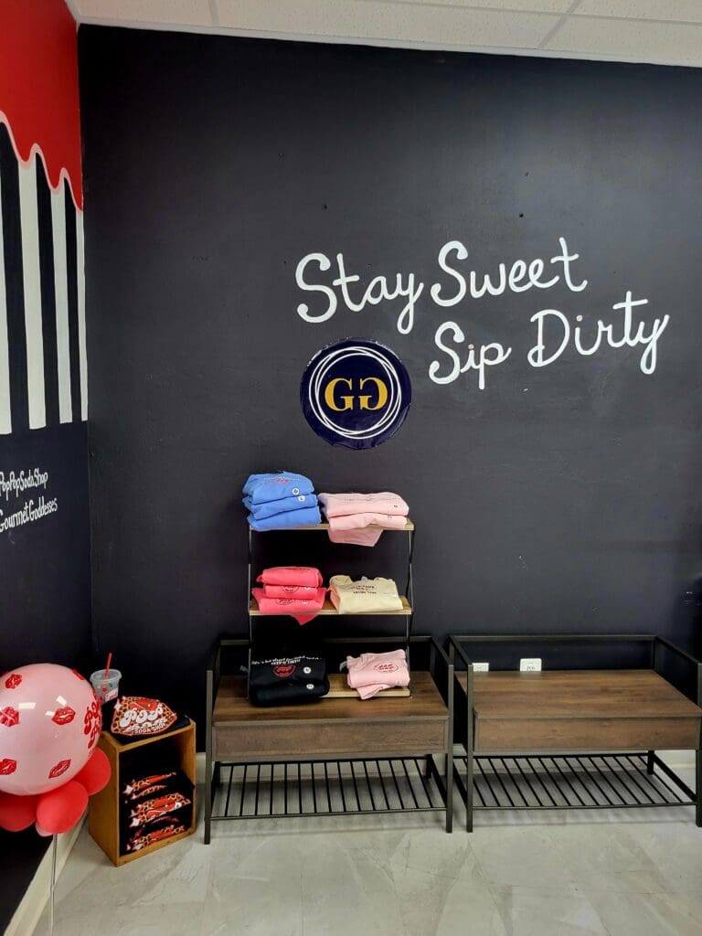 A cozy indoor beverage station with stacked colorful cups on metal shelving, decorated with a neon 'Stay Sweet Sip Dirty' sign on a dark wall.