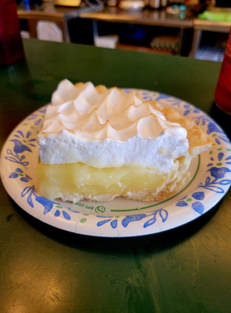 A slice of lemon meringue pie with golden custard filling and tall peaked white meringue topping sits on a decorative blue and white plate.