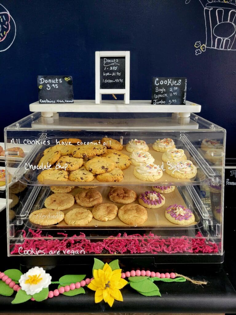 A bakery display case filled with golden donuts and pastries sits beneath a dark blue wall with decorative signage and colorful flower garland.
