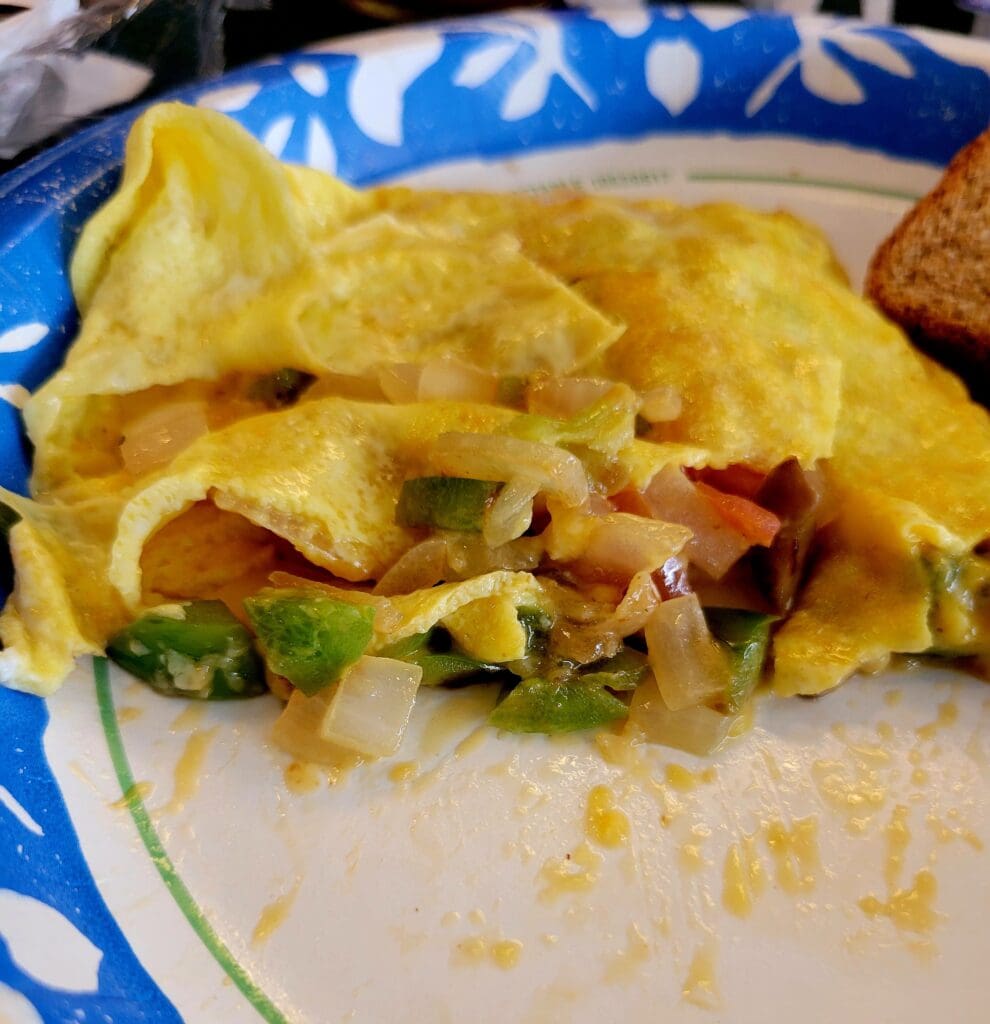 A close-up photograph of a golden omelet filled with lettuce, ham, and cheese served on a decorative blue and white plate with a slice of bread on the side.