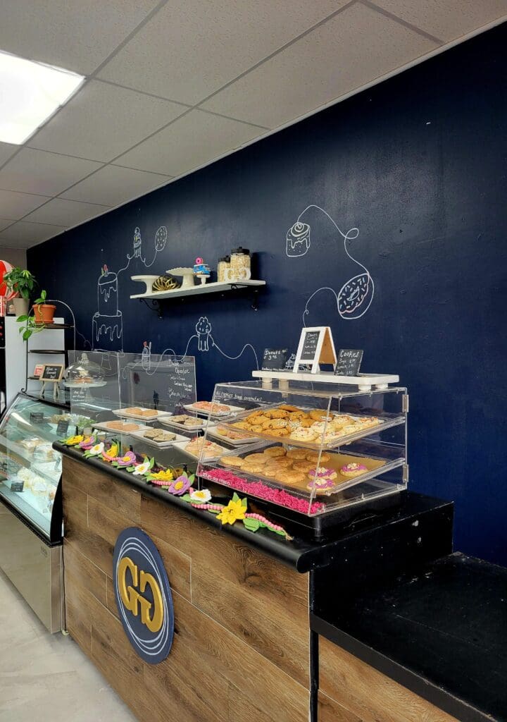 A modern bakery interior features a dark navy display counter with illuminated pastries and baked goods, complemented by navy walls with minimalist chalk drawings and wooden accents.