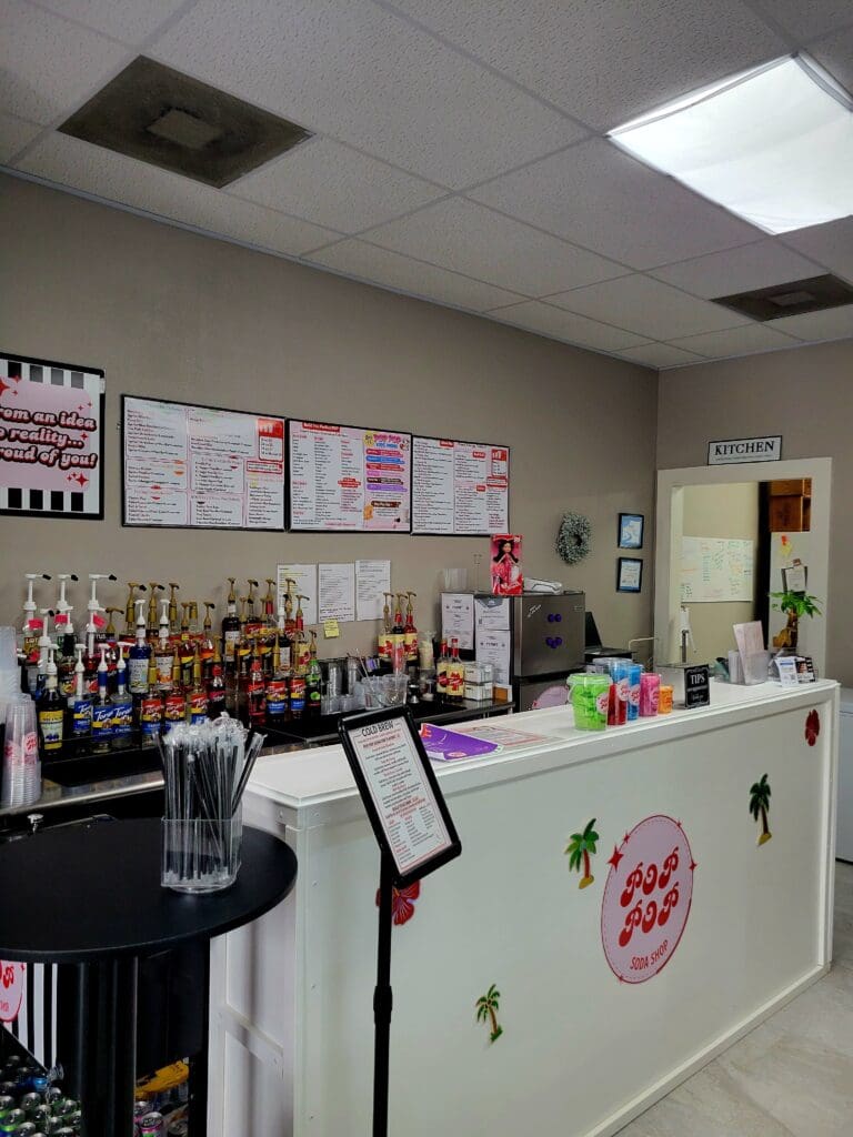 A small retail shop counter with product displays on shelves and walls, featuring a pink circular logo and various merchandise arranged for sale.