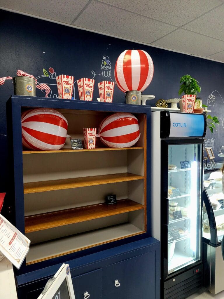 A carnival-themed concession stand display with red and white striped popcorn boxes, hot air balloon decorations, and a beverage cooler against a dark blue wall.