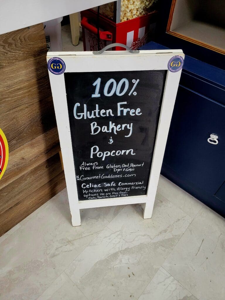 A white sandwich board sign advertising a 100% gluten-free bakery and popcorn stand sits on a concrete floor in an indoor retail or food service space.
