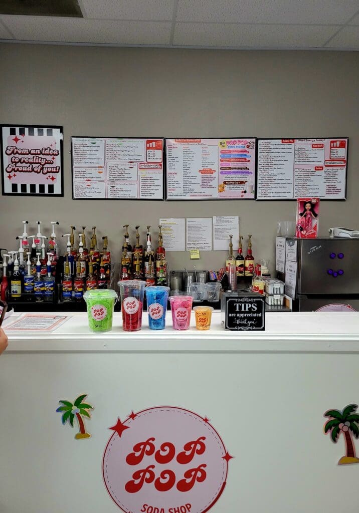 A coffee shop counter with colorful beverage dispensers, menu boards on the wall, and tropical palm tree decorations displayed below.