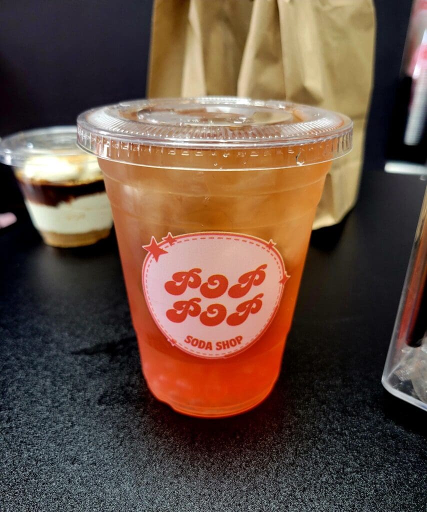 A clear plastic cup of amber-colored beverage with a branded logo sits on a dark counter with additional drinks visible in the background.