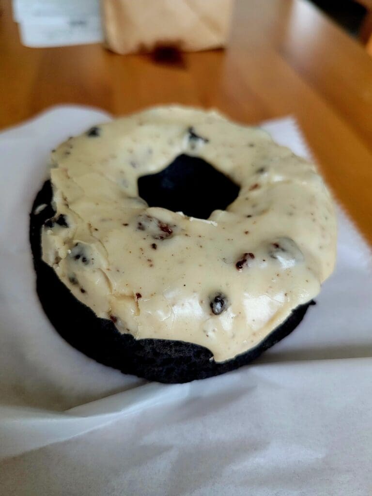 A close-up shot of a chocolate-frosted donut with white icing and dark chocolate drizzle on a white plate resting on a wooden table.