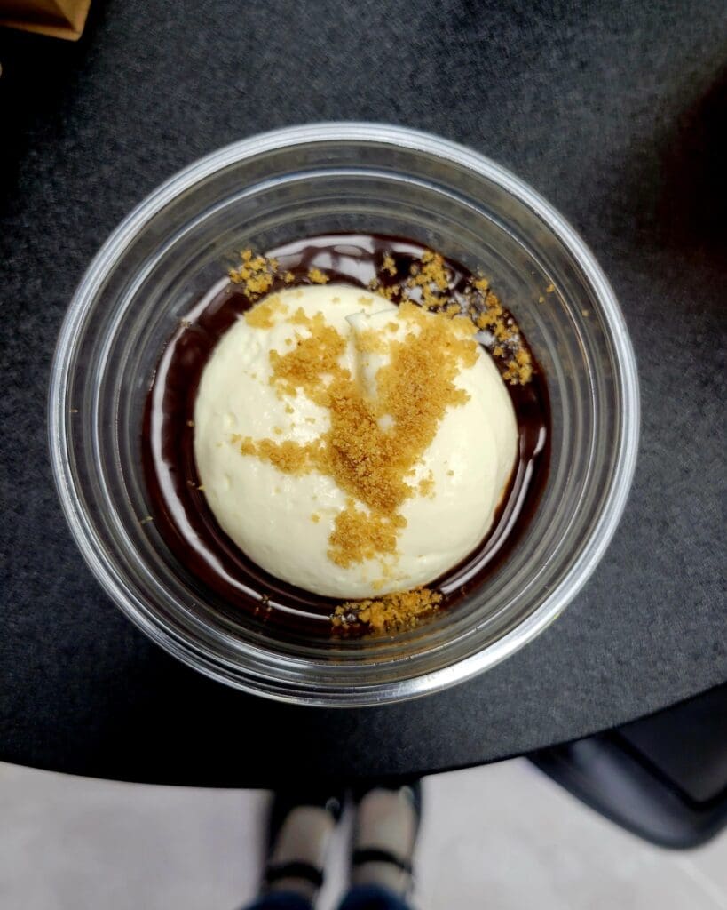 A close-up overhead view of a creamy dessert topped with golden-brown sugar or spice in a clear glass bowl against a dark textured surface.