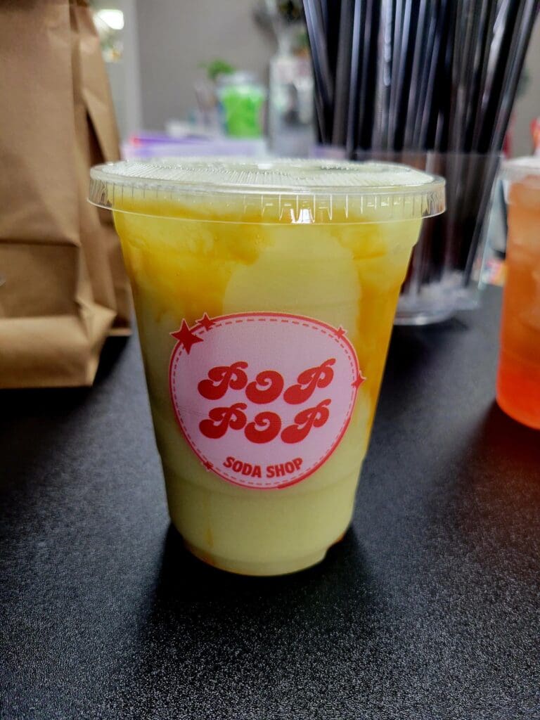 A close-up shot of a yellow-green bubble tea drink with a branded cup label and clear plastic lid, sitting on a dark counter with a paper bag and storefront visible in the soft-focused background.