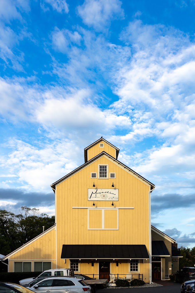 A bright yellow barn with a peaked roof stands prominently against a blue sky with white clouds, with parked vehicles visible at its base.