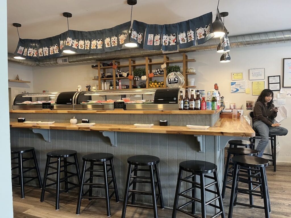 A modern bar or restaurant interior featuring a wooden counter with black bar stools, pendant lighting, and shelved beverages, with one person visible in the background.