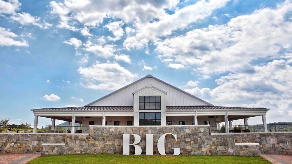 A modern agricultural event venue with a large barn-style building featuring stone walls and covered porches, with prominent 'BIG' lettering in the foreground under a bright blue sky with scattered clouds.