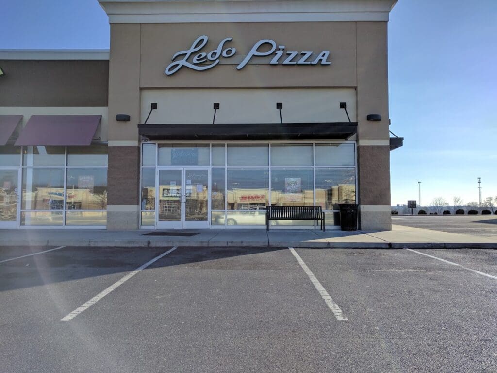 A closed or quiet pizza restaurant storefront with a tan and gray exterior, empty parking lot, and clear signage on a bright daytime.