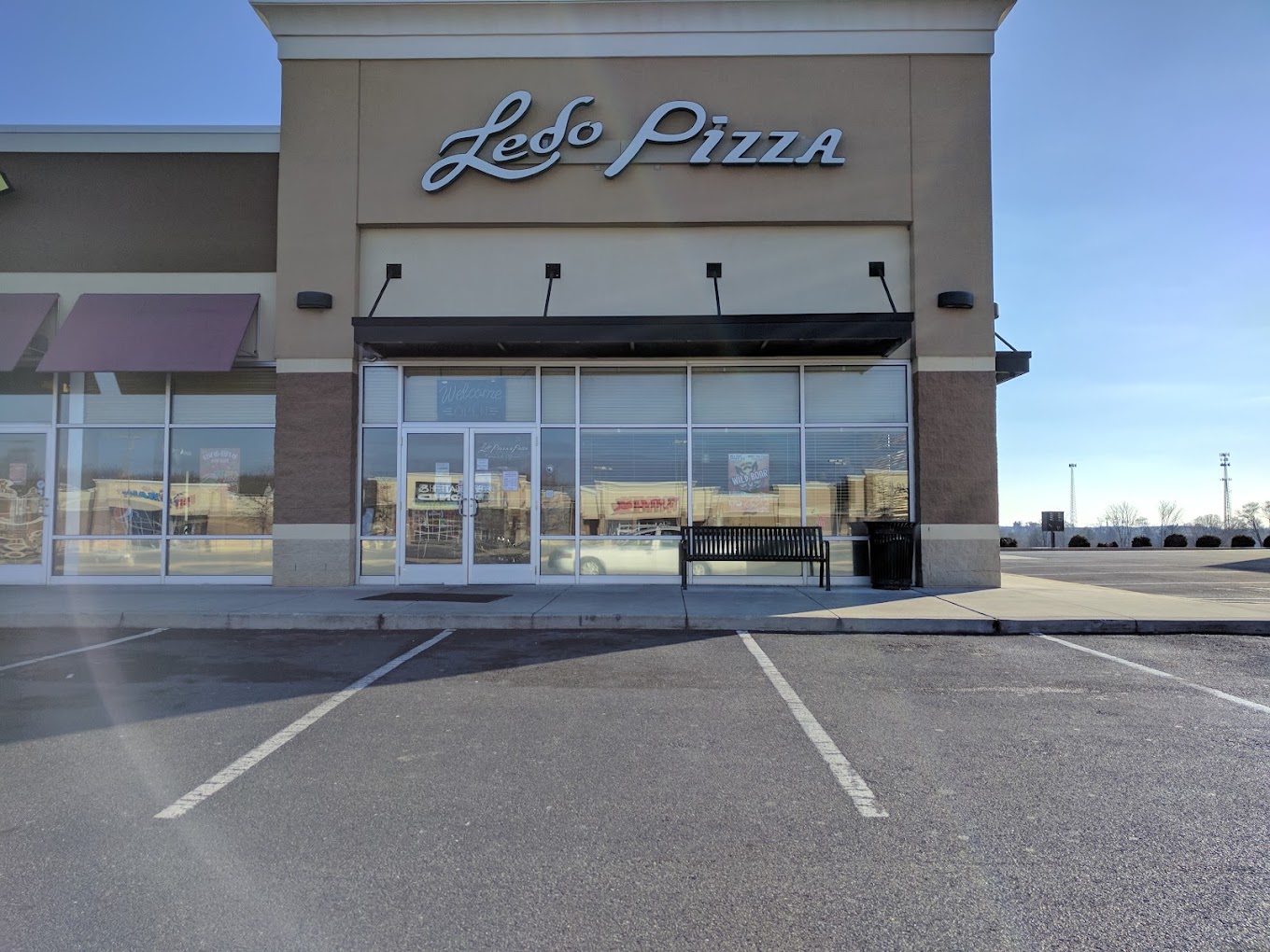 A closed or quiet pizza restaurant storefront with a tan and gray exterior, empty parking lot, and clear signage on a bright daytime.