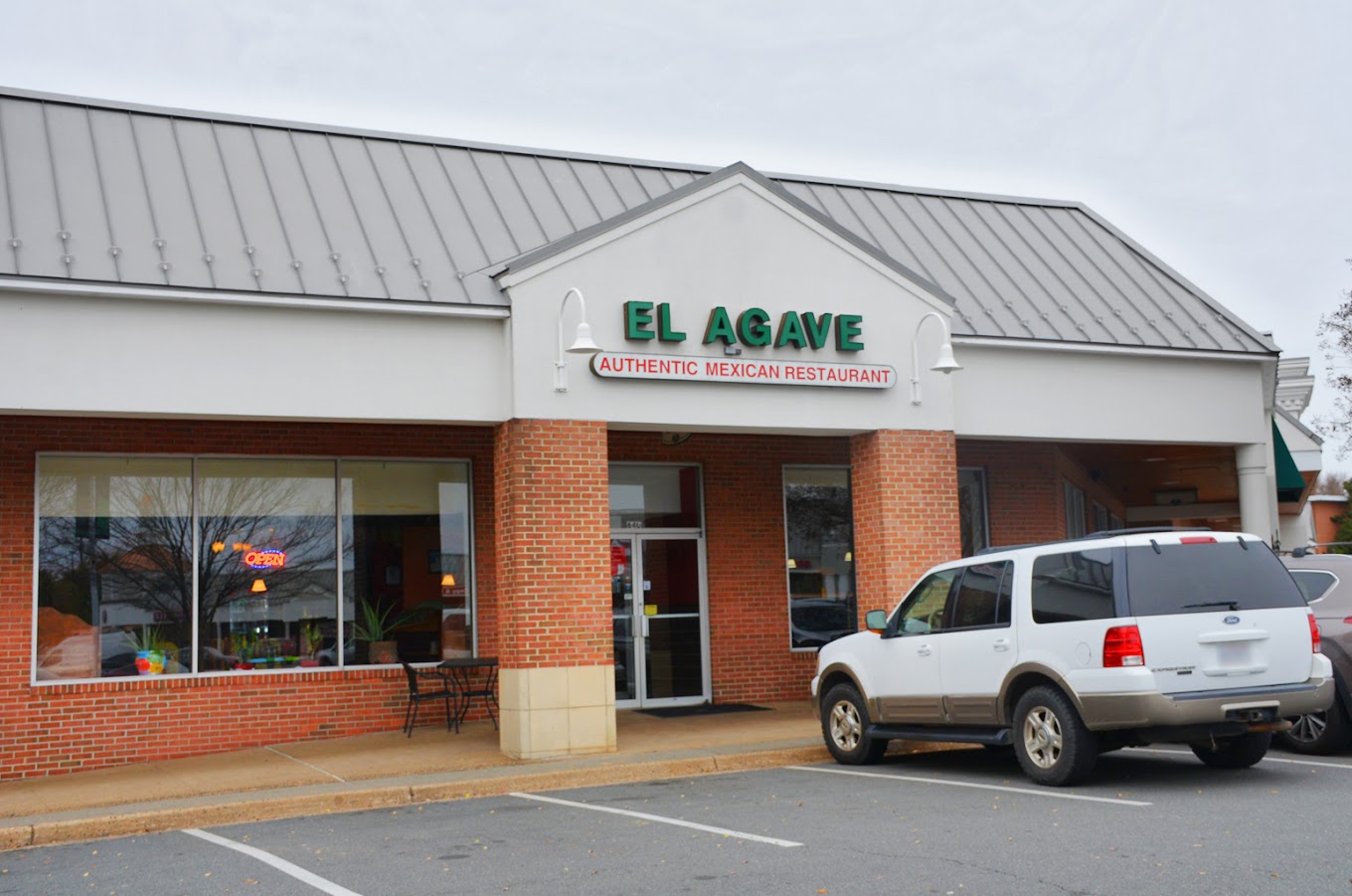 A daytime photograph of El Agave restaurant's storefront with brick and metal siding, featuring a parked silver vehicle in an empty parking lot.