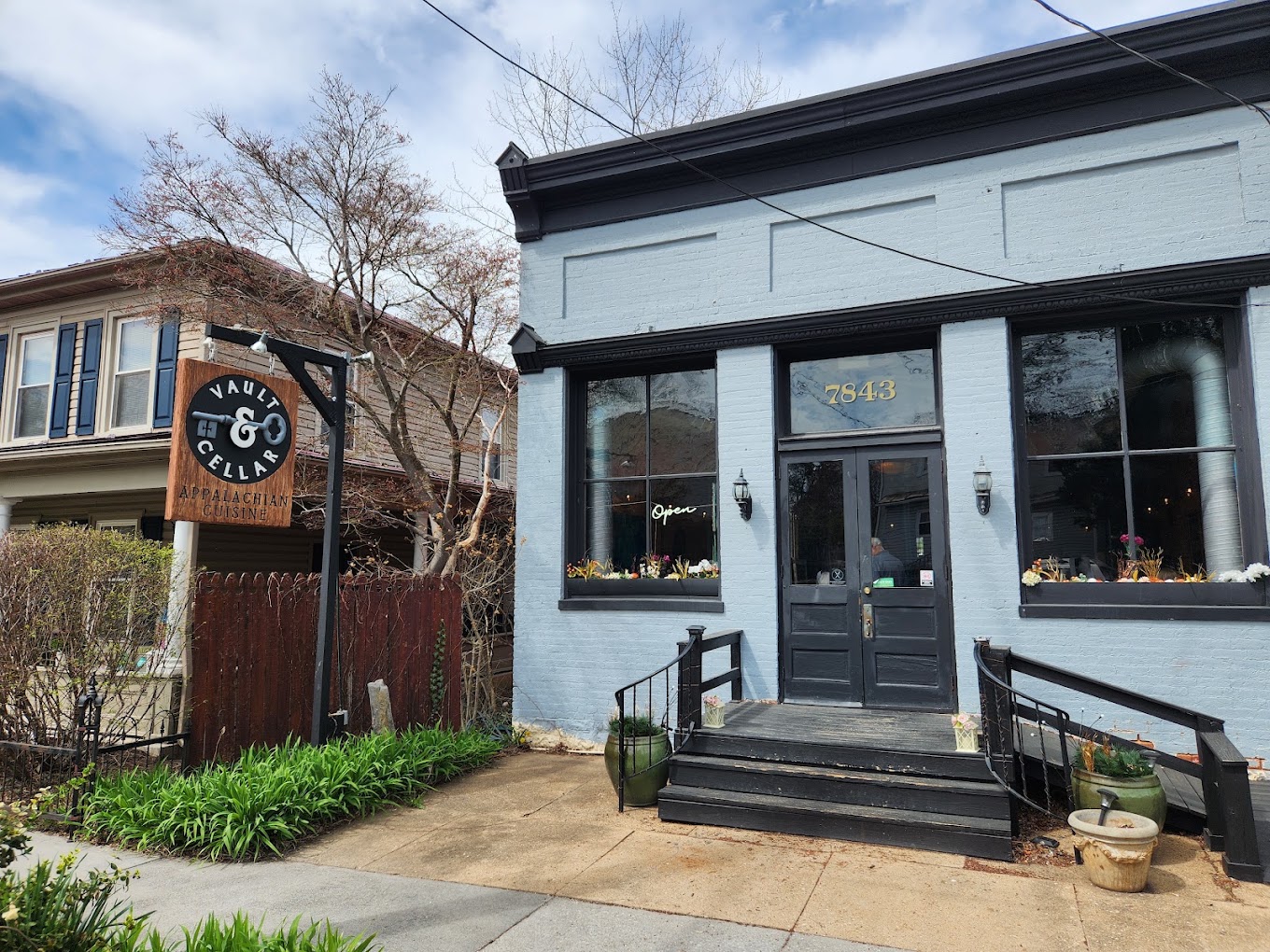 A charming contemporary storefront with modern dark-trimmed windows, a prominent wooden clock sign, and tasteful landscaping on a quiet day.