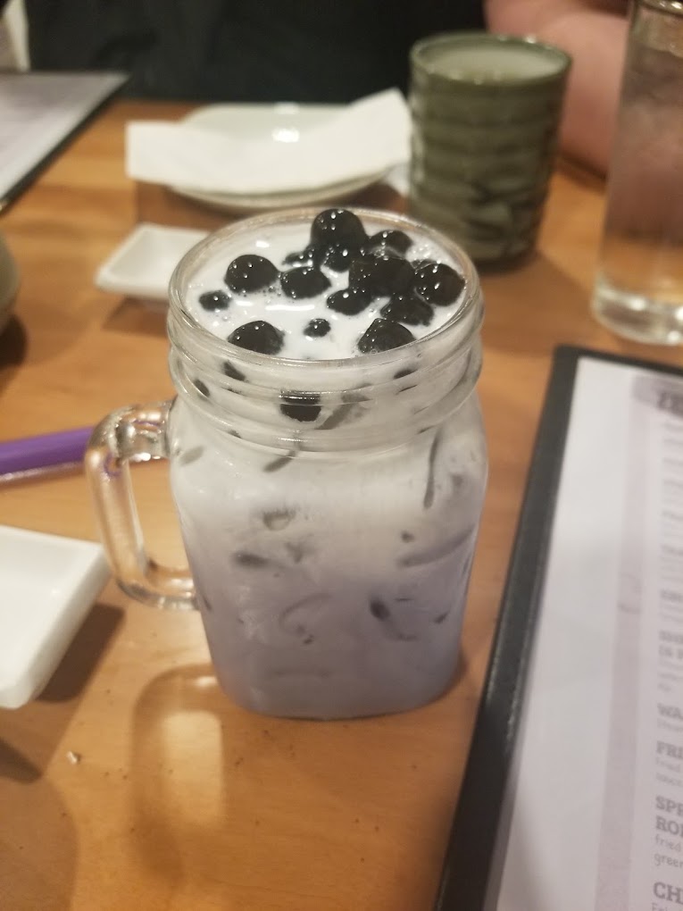 A close-up of a refreshing boba tea drink with dark tapioca pearls and whipped cream topping, served in a clear glass on a wooden table in an indoor cafΓ© setting.