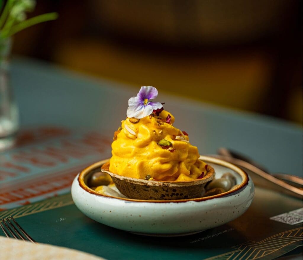 A carefully plated dessert featuring bright yellow shaved mango ice topped with a delicate purple flower sits in a white ceramic bowl on a decorative patterned surface.