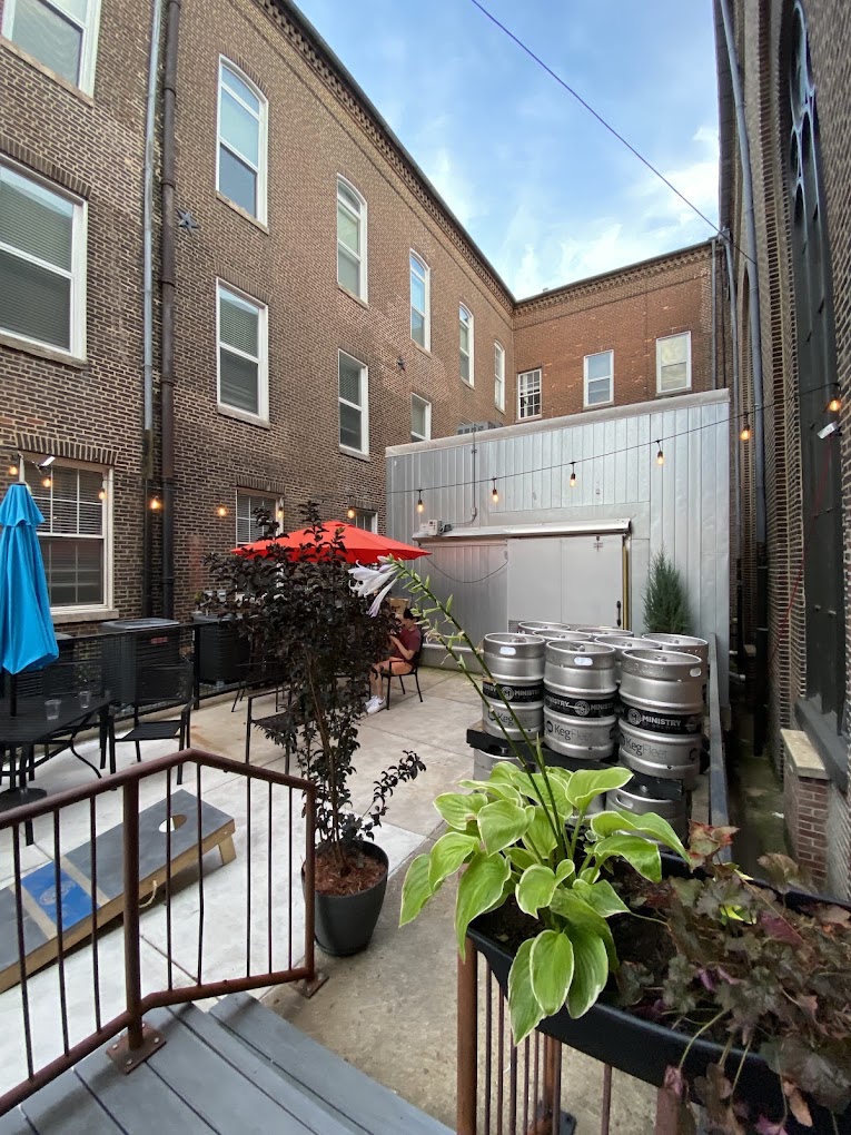 A quiet urban courtyard between brick buildings features potted plants, beer kegs, and outdoor seating in what appears to be a restaurant or bar venue space.