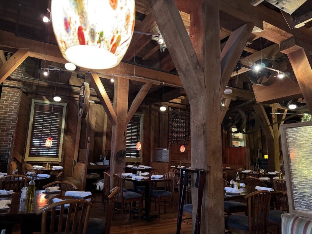 An upscale restaurant interior featuring rustic wooden beam architecture, warm pendant lighting, and multiple dining tables set for service.