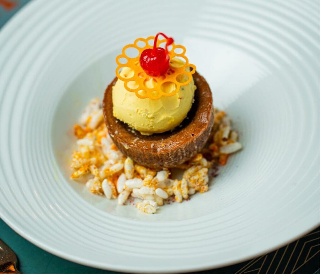 An elegantly plated gourmet dessert featuring a layered chocolate and vanilla creation topped with a yellow mousse dome, macaroon, and red cherry garnish, presented on a white ribbed plate.