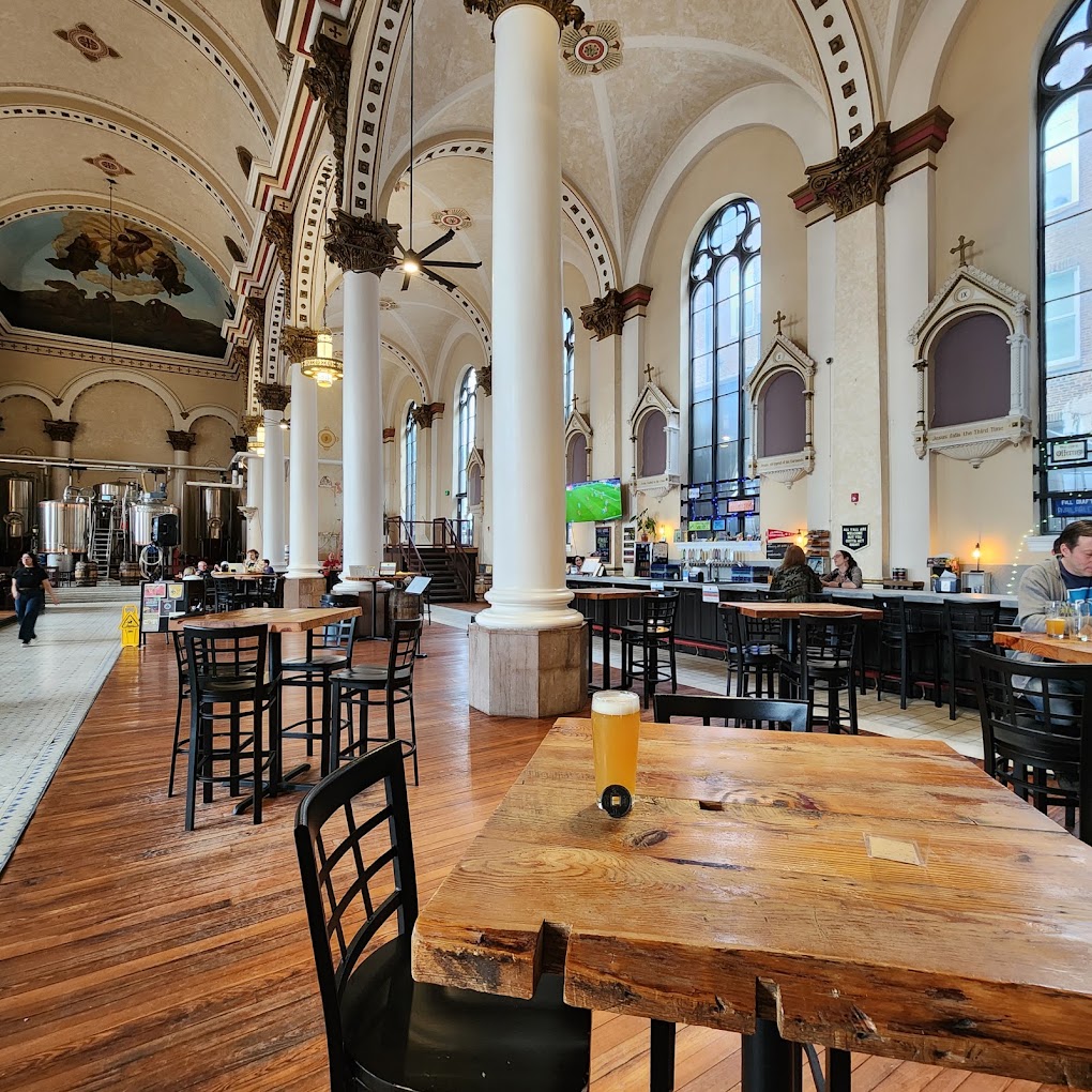 An elegant restaurant or event venue with soaring ceilings, decorative columns, wooden tables, and black chairs arranged throughout a spacious interior with classical architectural details.