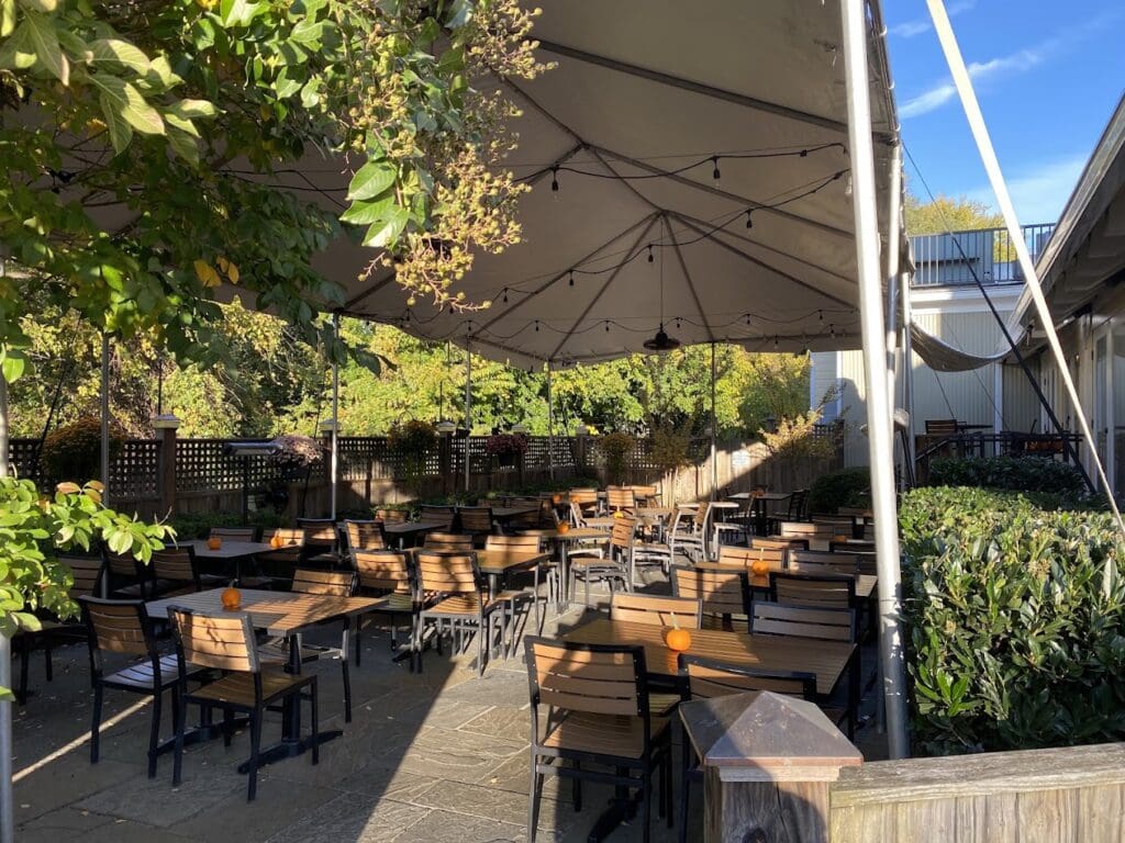 An inviting outdoor patio dining area with wooden tables and chairs arranged under a large shade structure, surrounded by lush greenery and climbing vines on a clear sunny day.