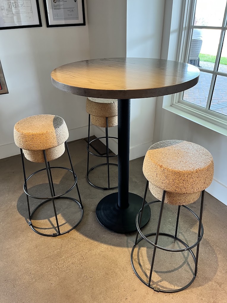 A modern bar-height table with two cork-topped stools in an interior space with natural light from large windows.