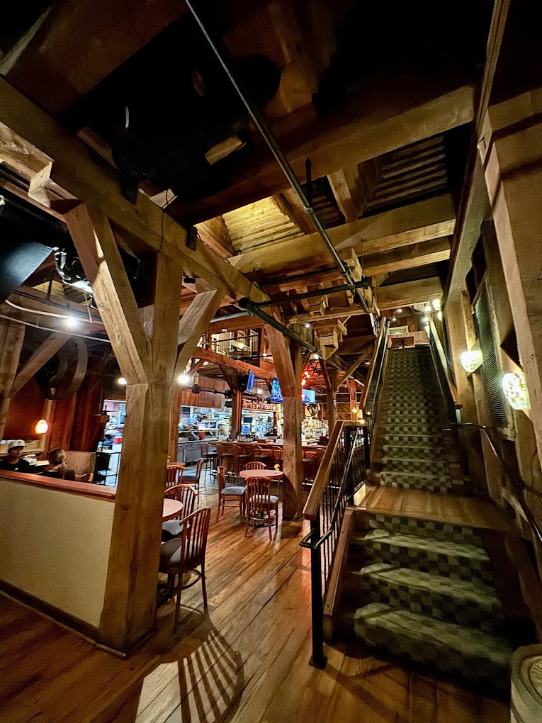 A rustic interior shop or small venue illuminated by warm lighting, featuring exposed wooden ceiling beams, shelving with merchandise, and stairs, creating a vintage or antique store atmosphere.