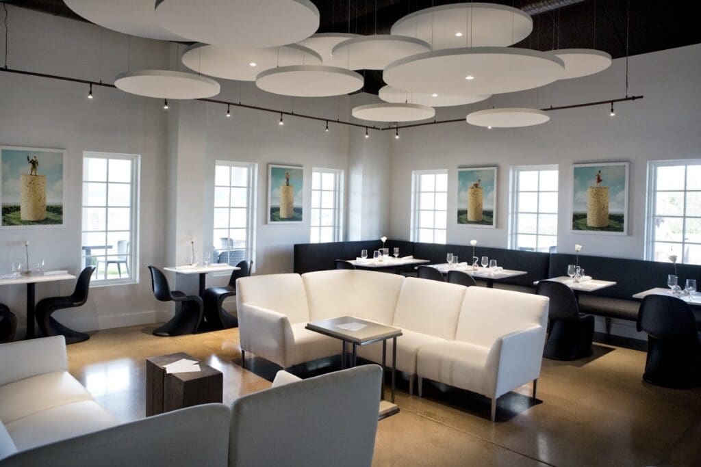 A modern, minimalist lounge or hospitality space features contemporary furniture, geometric ceiling lighting, and natural light from multiple windows.