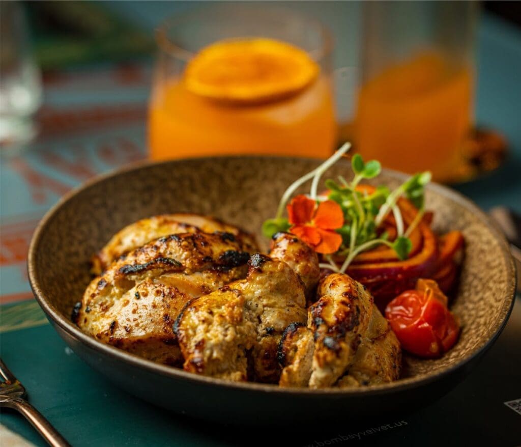 A close-up of grilled meat skewers served in a dark bowl with roasted vegetables and garnish on a teal dining surface, with beverages in the background.