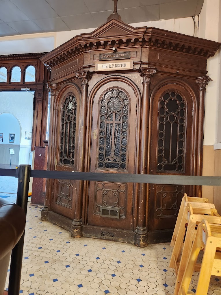 A wooden confessional booth with ornate Victorian architectural details and stained glass windows displayed indoors, likely in a museum or restoration workshop.