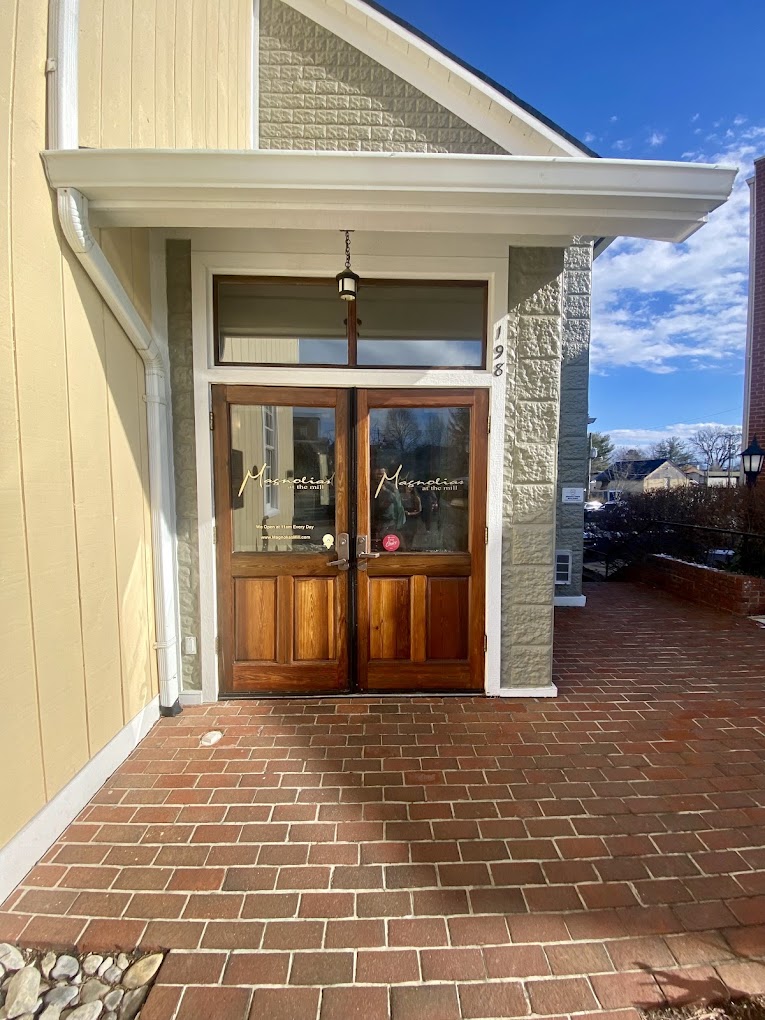 A residential home entrance features wooden double doors with glass panes, set within white trim and stone accents, opening onto a brick patio under clear blue sky.
