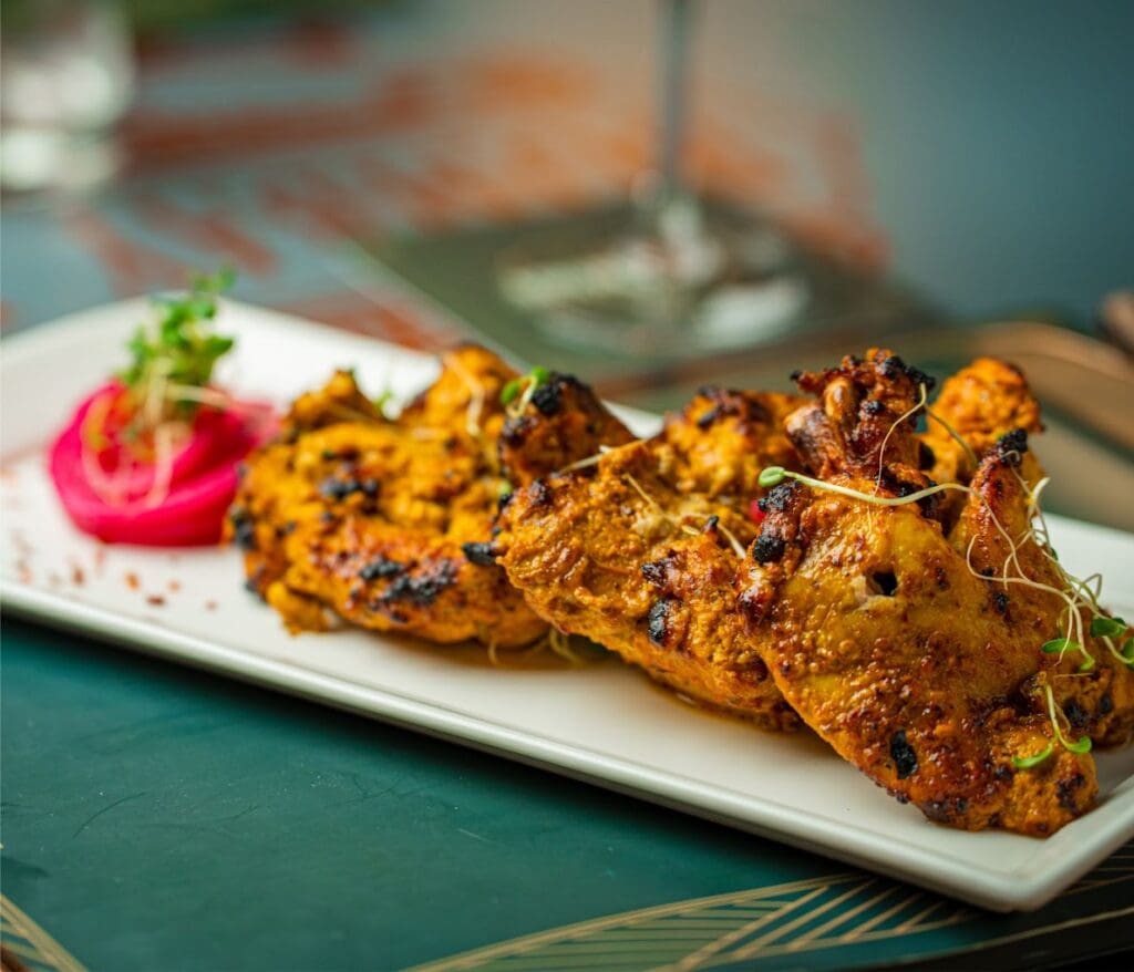 Beautifully plated grilled chicken skewers with charred exterior, garnished with fresh herbs and pink radish slices on a white plate against a teal background.