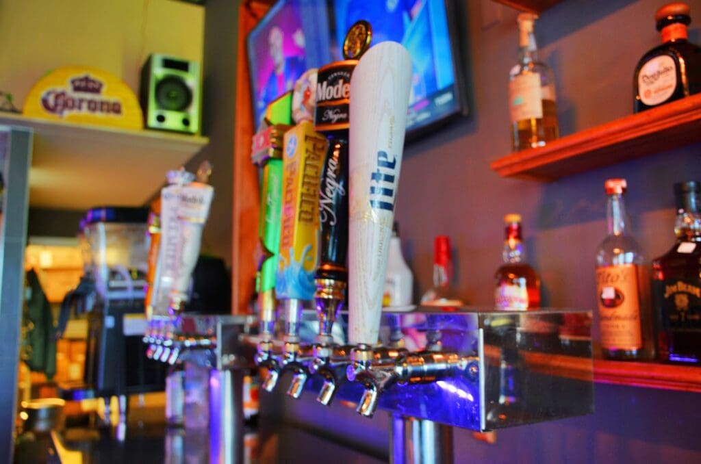 A colorful bar interior showcases multiple beer taps and illuminated liquor bottles with vibrant purple and blue neon lighting.