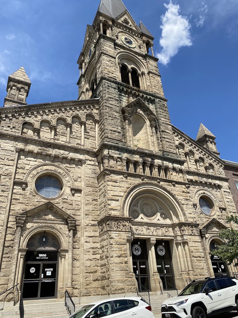 A grand stone church building with a prominent bell tower and ornate Romanesque architecture stands against a bright blue sky.