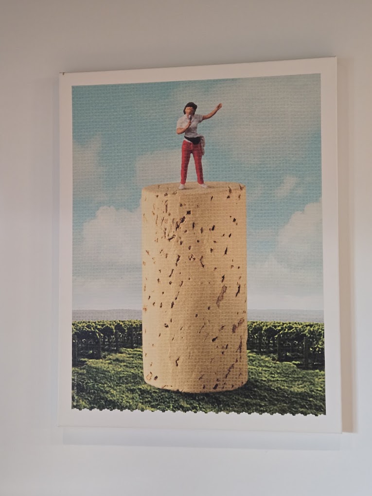 A surreal artwork depicting a solitary figure standing atop a massive cork cylinder in an expansive landscape under a partly cloudy sky.