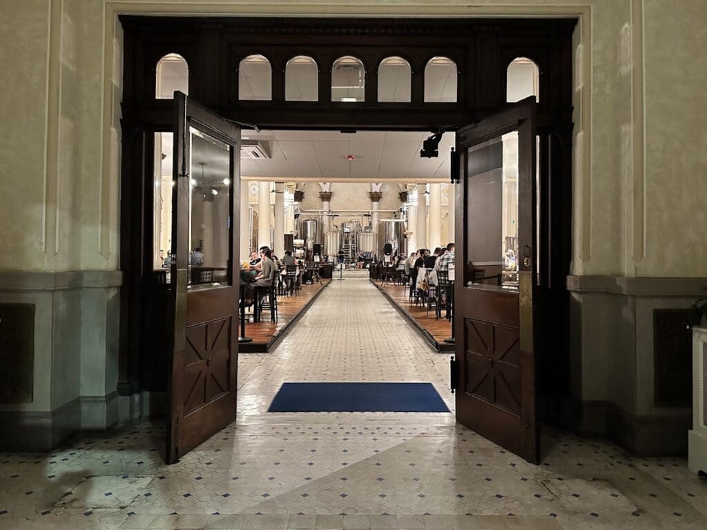 A symmetrical view through grand wooden doors into a formal institutional hallway with classical architecture, arched windows, and sparse visitors walking through.