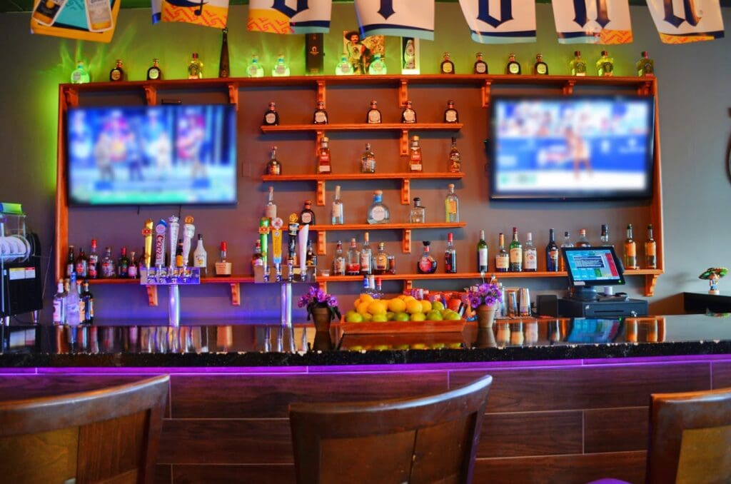 A well-lit bar interior features an illuminated back counter with multiple liquor bottles, TV screens, and purple ambient lighting creating a warm, inviting atmosphere.