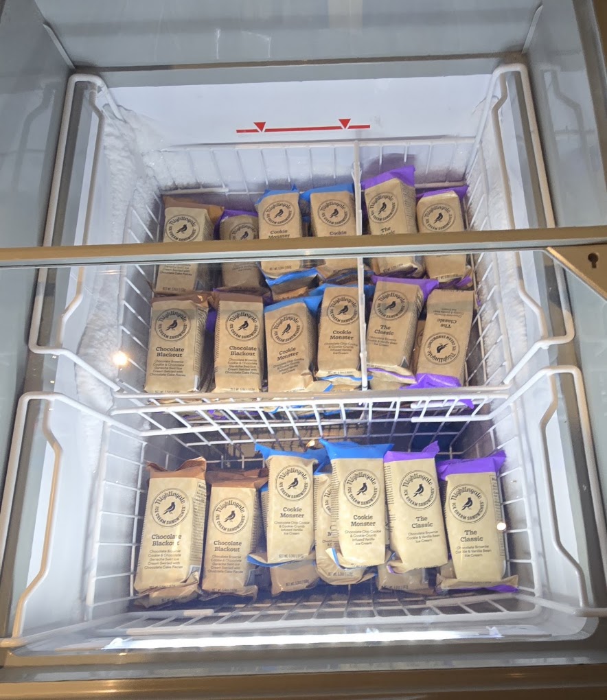 An overhead view of an organized freezer with multiple wire baskets containing kraft cardboard boxes with blue labels, appearing to be a storage or inventory system.