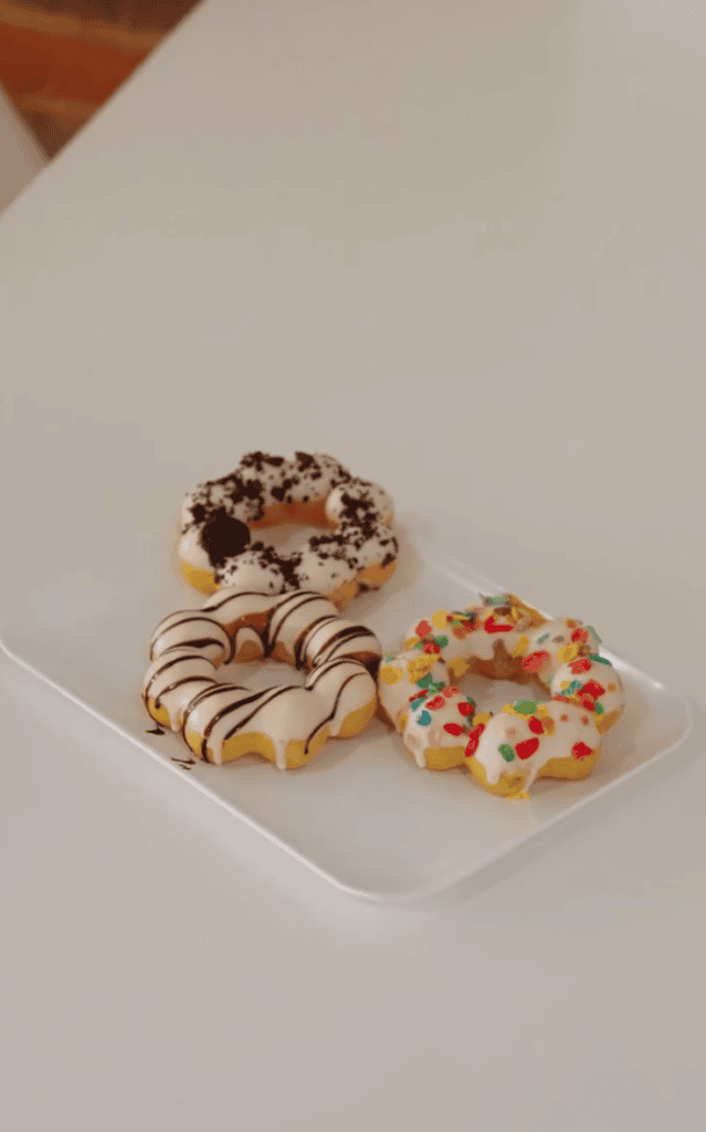 Three decorated donuts with chocolate drizzle, cookie crumbs, and colorful sprinkles sit on a white tray against a white surface.