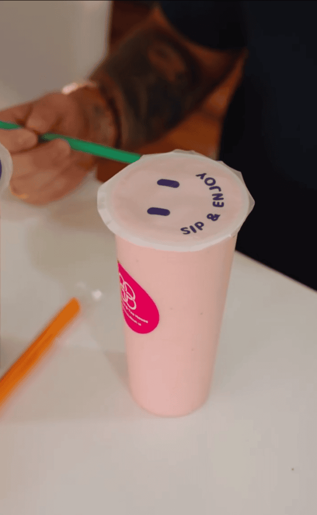 A pink smoothie cup with a 'Sip & Enjoy' lid sits on a table while a person holds a green straw in the background.