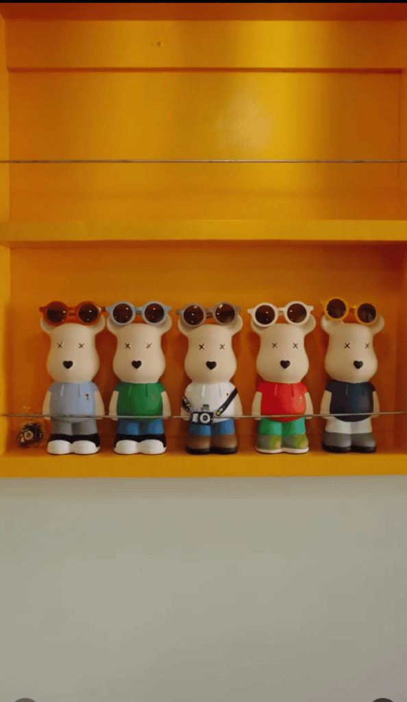 Five identical bear figurines wearing different colored sunglasses and outfits are displayed in a row on a bright orange shelf.