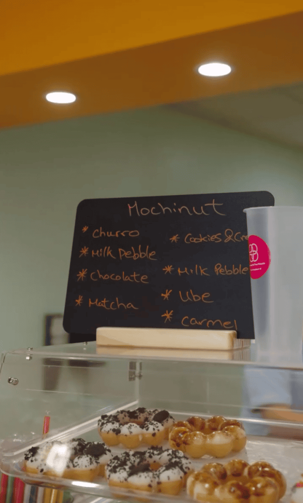 A chalkboard menu listing Mochinut flavors sits above a display case of decorated mochi donuts in a bakery.